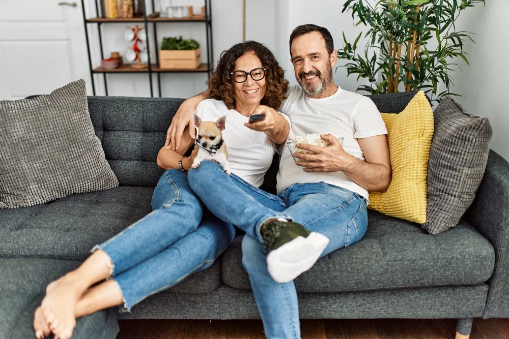 Man and woman with chihuahua on couch watching tv.